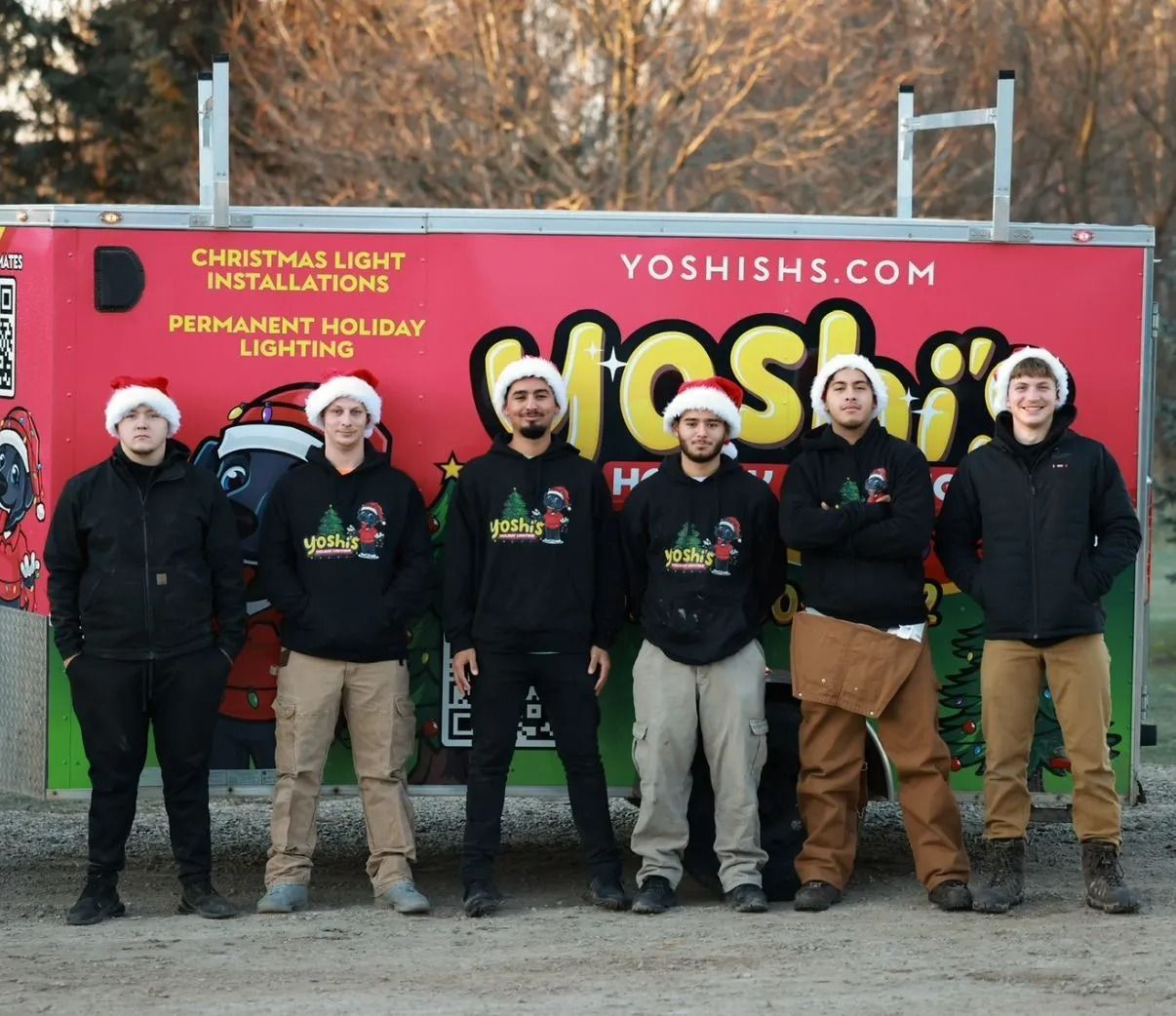 Six people wearing Santa hats stand in front of a trailer that advertises Christmas light installation.
