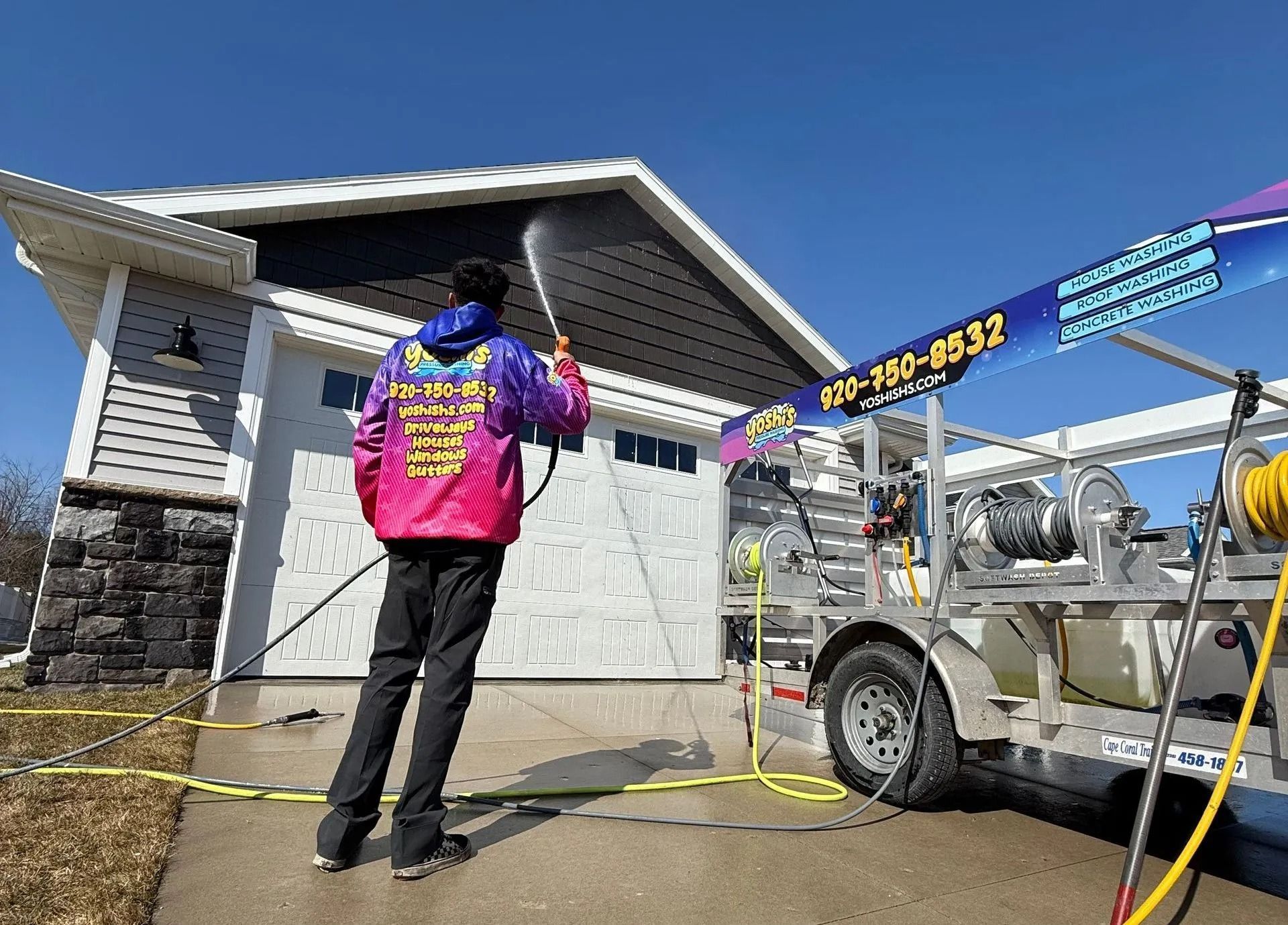 Person power washing the exterior of a house with a pressure washer; blue sky overhead.