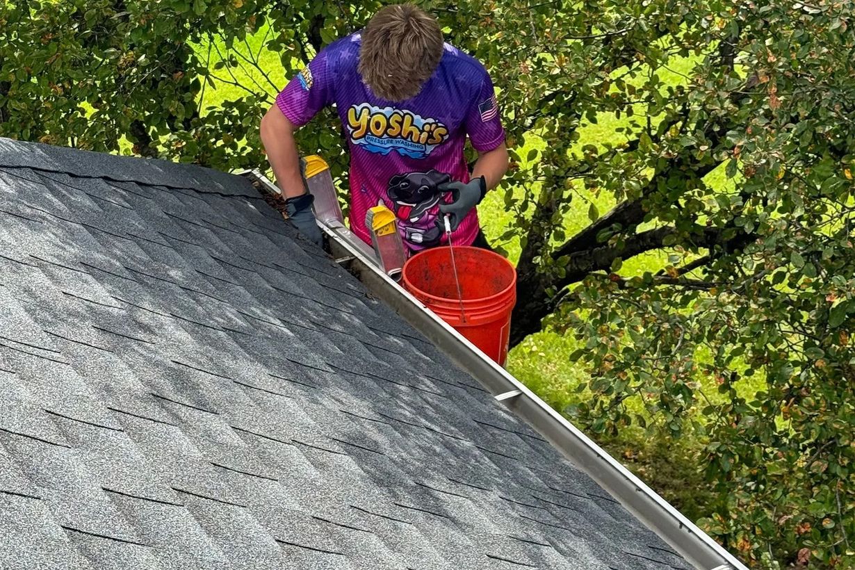 Person on a roof cleaning a gutter with a bucket, wearing a colorful shirt, on a sunny day.