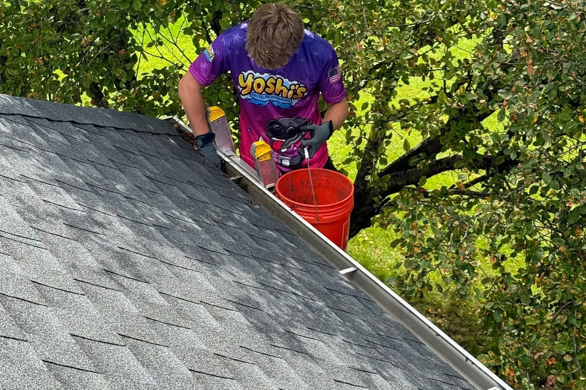 Person on a roof cleaning a gutter with a bucket, wearing a colorful shirt, on a sunny day.