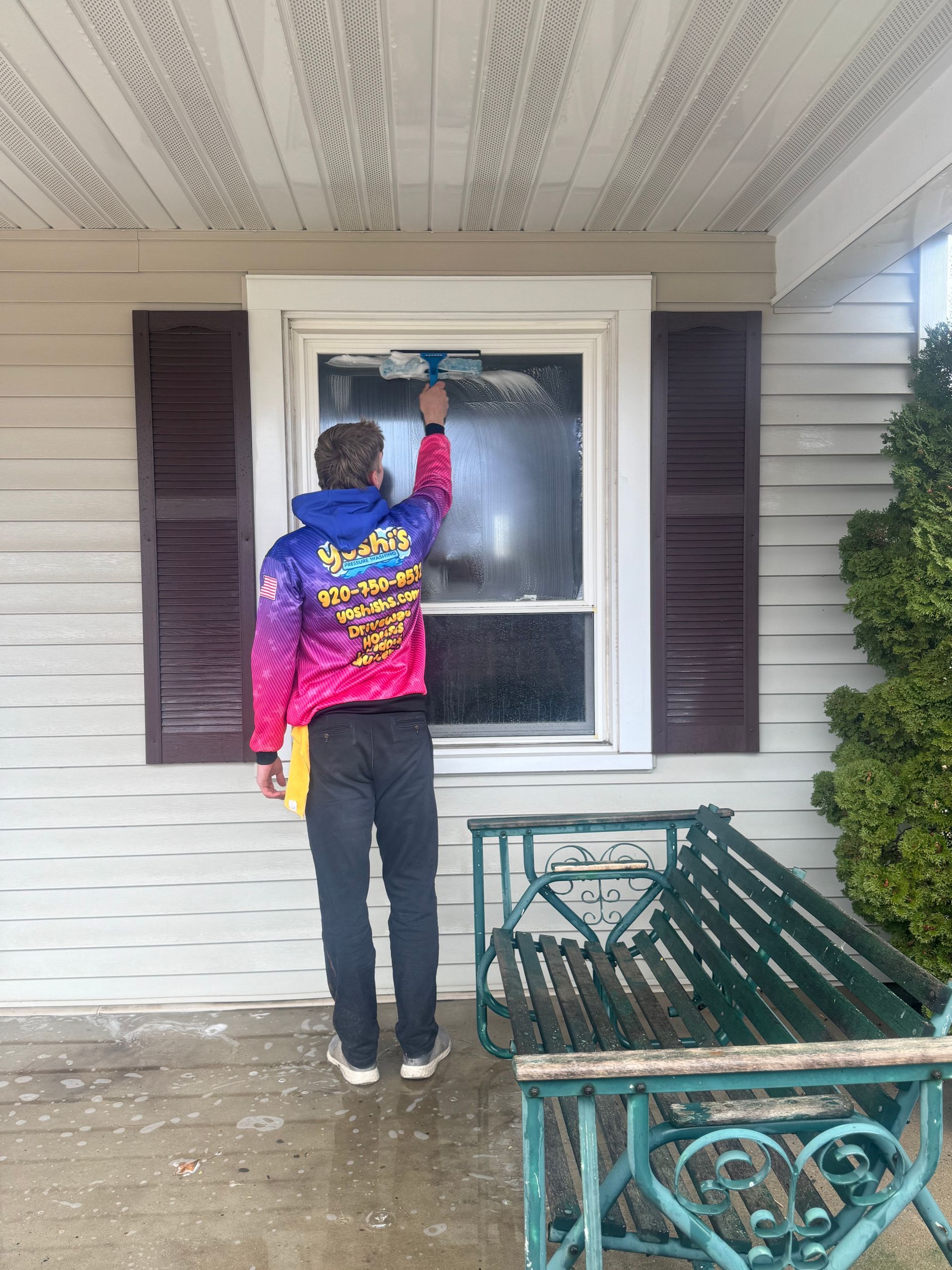 Person cleaning a window with a squeegee on a porch. They wear a colorful jacket and dark pants.