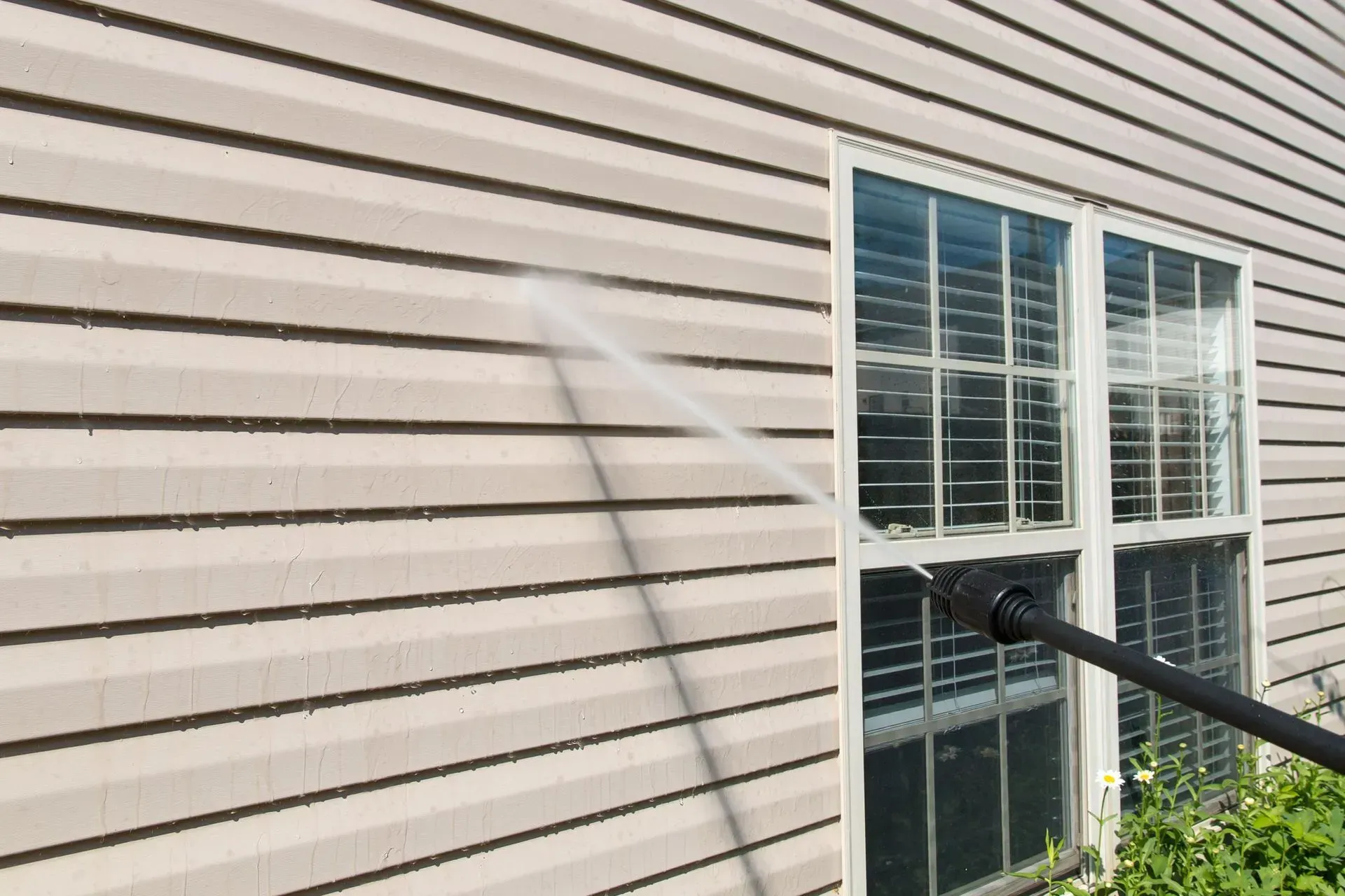 A pressure washer spray cleans light beige vinyl siding next to a white-framed window on the exterior of a house.
