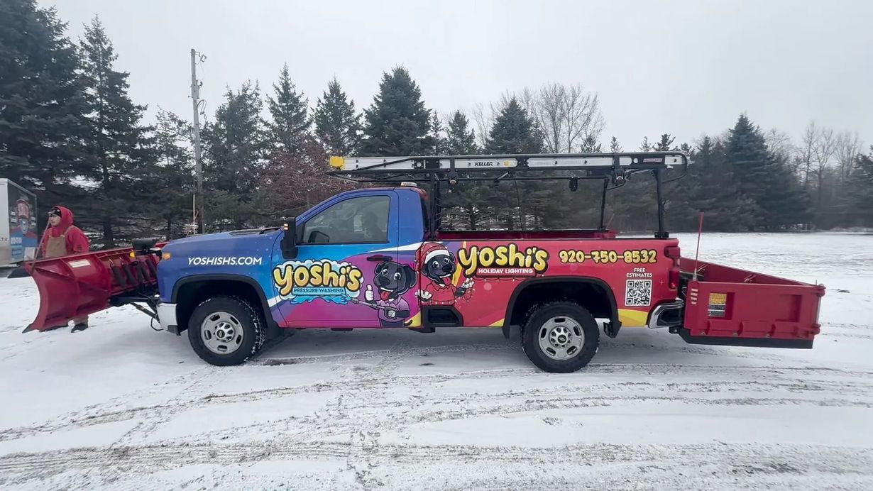 Truck with snowplow and Yoshi's logo plowing snow, outdoors.