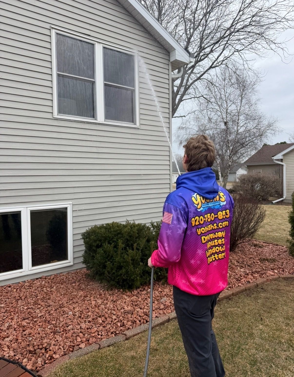 Person spraying a house, wearing a colorful jacket. House has siding and windows, yard with bushes and mulch.