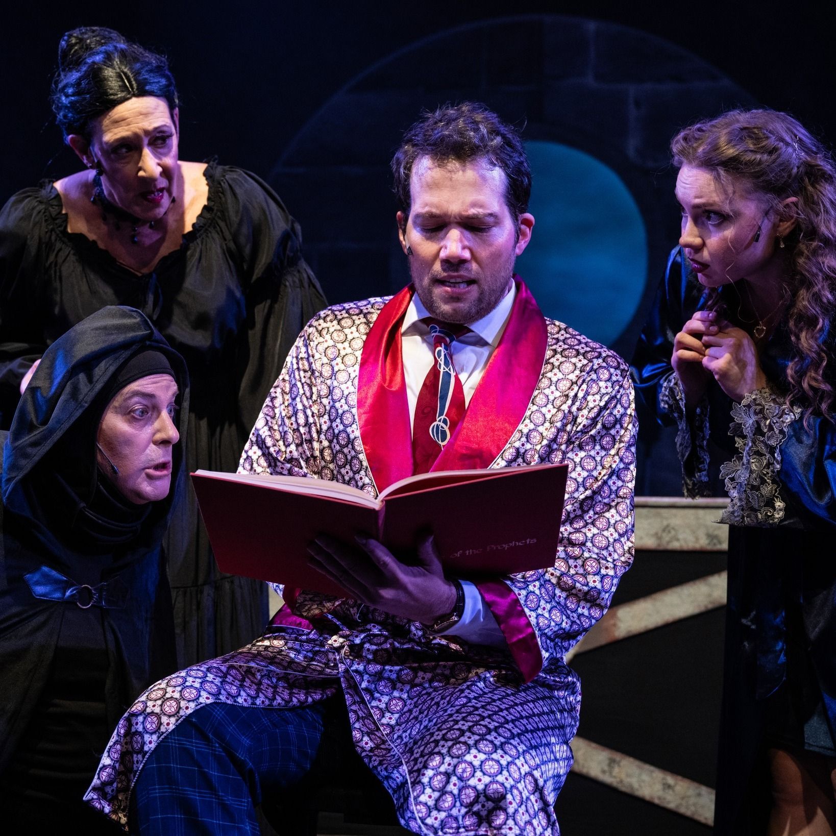 A man in a robe is reading a book while three women look on.