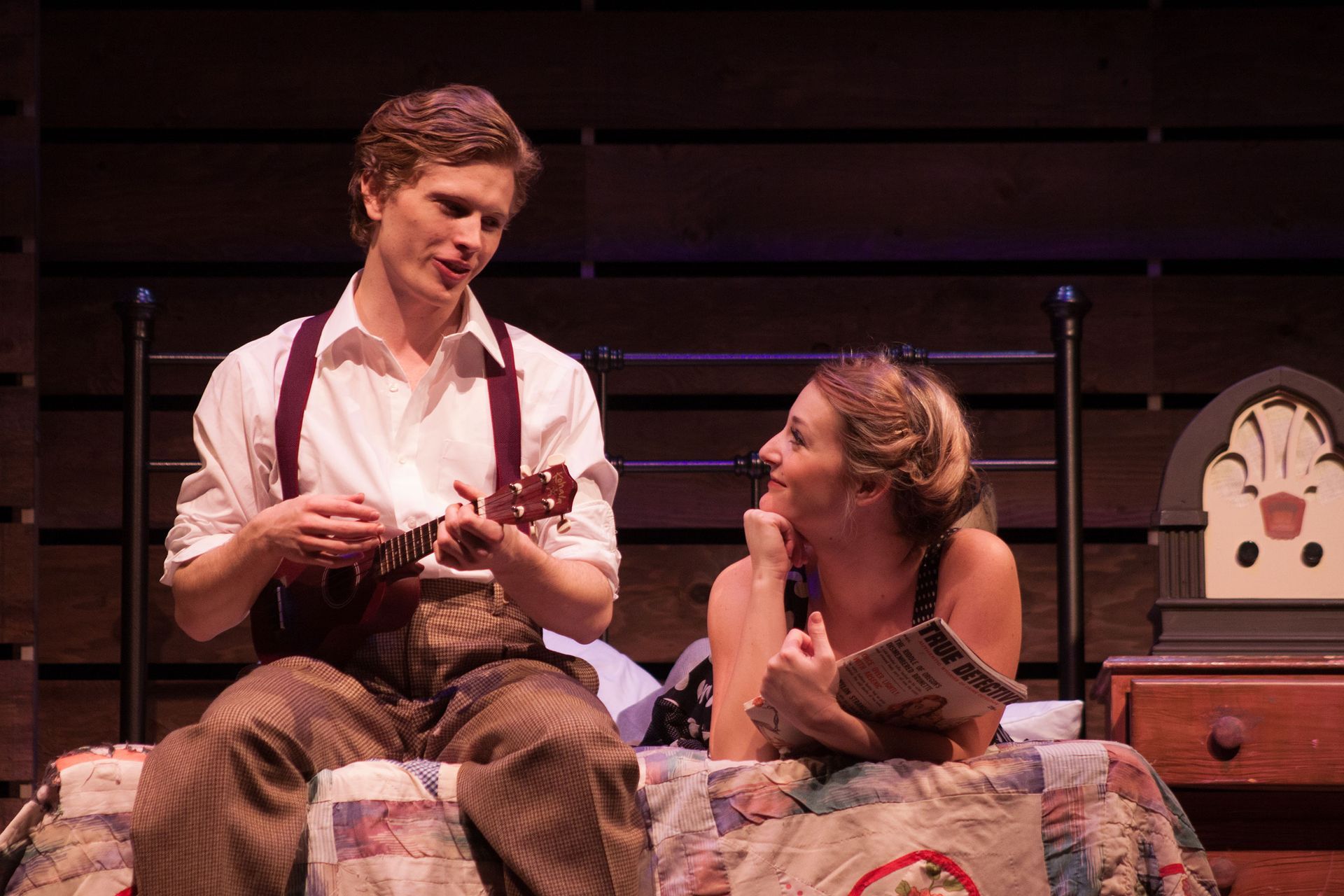 A man is playing a guitar and a woman is sitting on a bed.