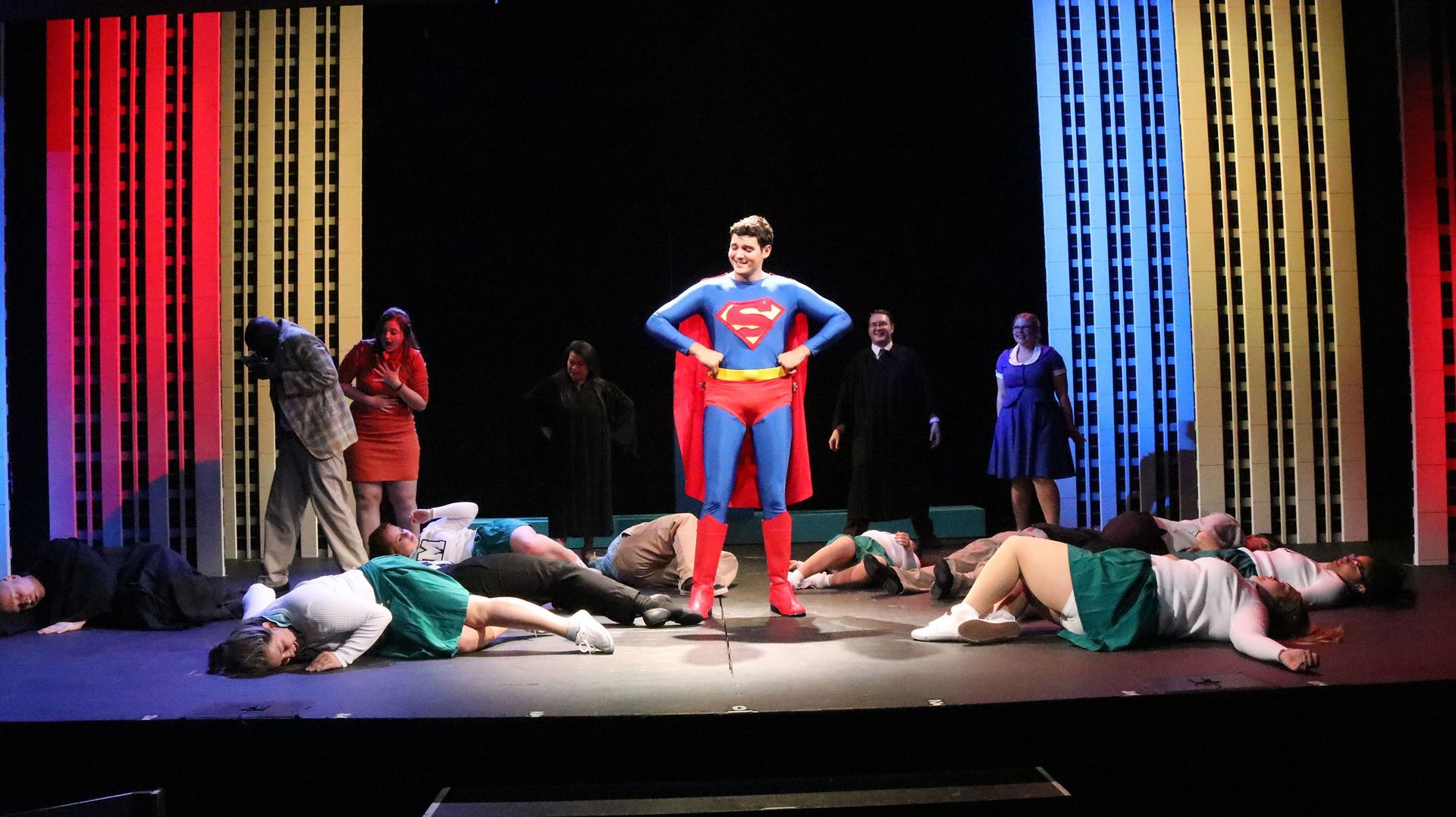 A man in a superman costume is standing on a stage surrounded by people laying on the floor.