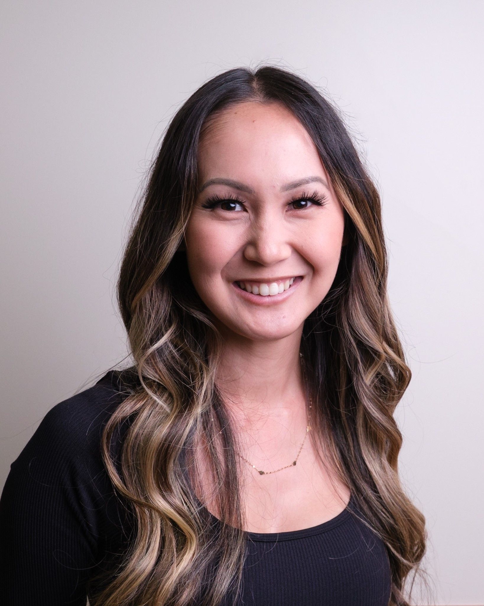 Woman with long, wavy brown hair smiles at the camera, wearing a black top, set against a light gray background.