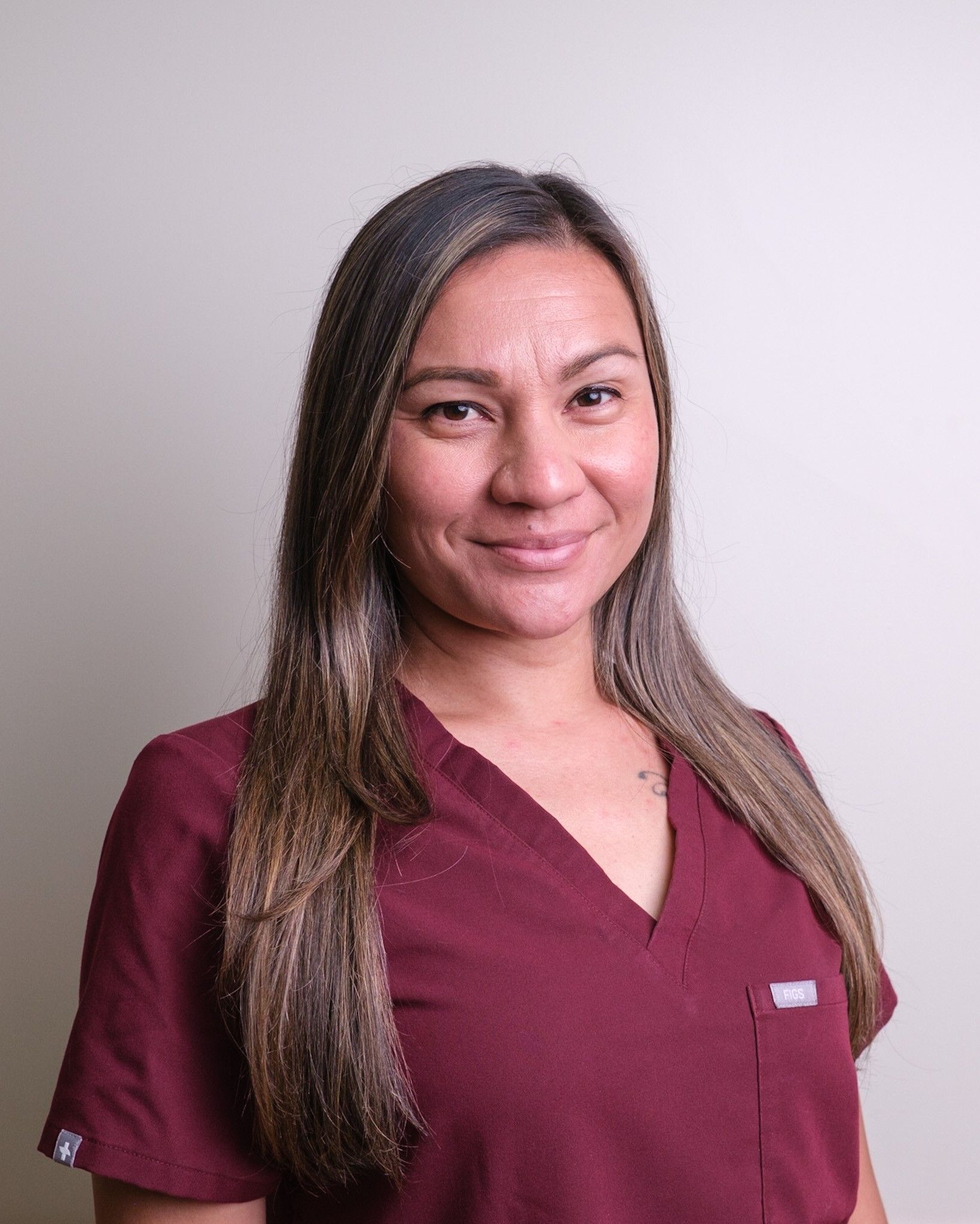Woman with long brown hair wearing a burgundy scrub top, smiles in front of a plain wall.