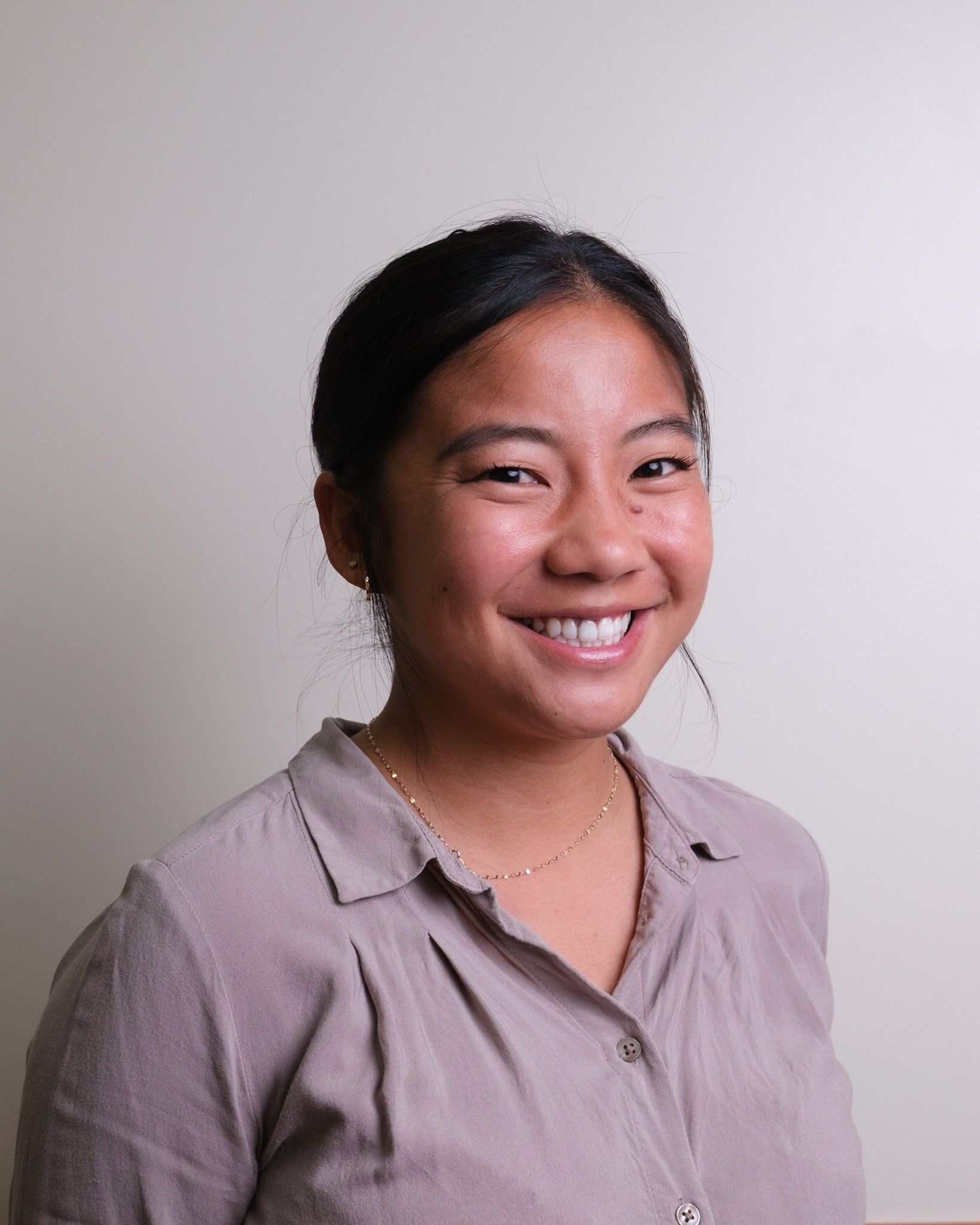 Young woman with dark hair smiling, wearing a tan button-up shirt, against a light background.