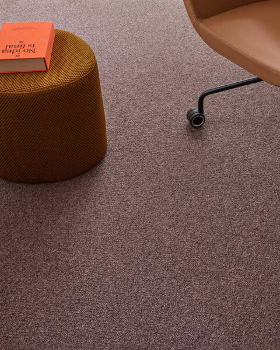 Mustard-colored ottoman with a book, part of an office chair, and textured brown carpet.