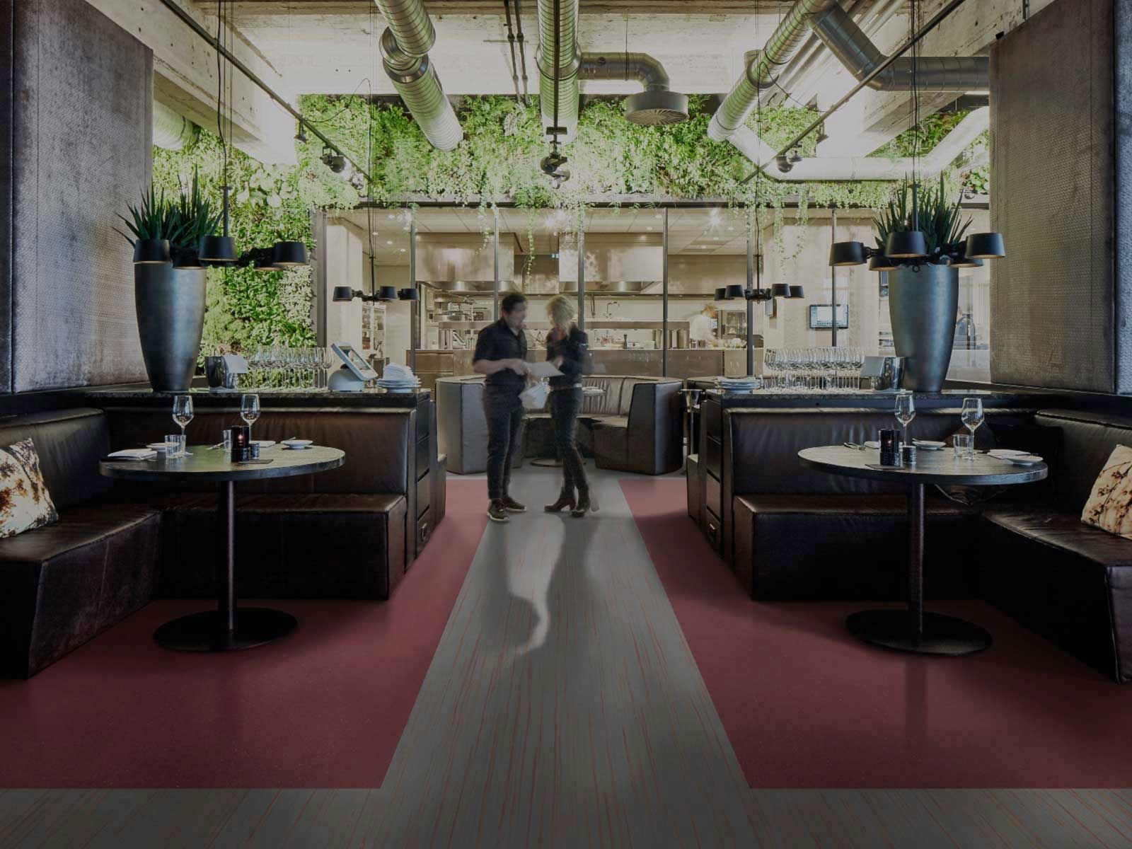 Restaurant interior: couple conversing near booths, pink and gray floor, greenery wall, industrial ceiling.