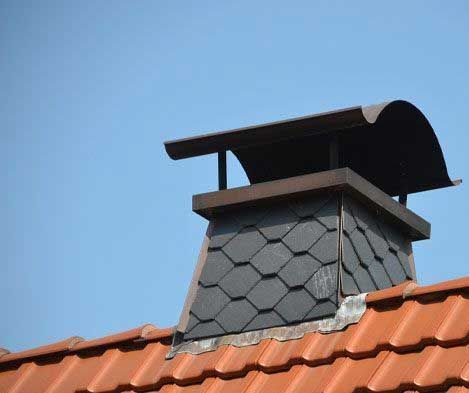 Chimney on a red-tiled roof against a clear blue sky, featuring a slate-like finish and a metal rain cap.
