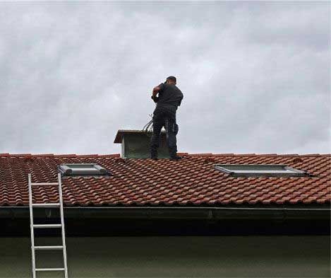 A person stands atop a roof near a chimney, appearing to be working. A ladder leans against the roof.