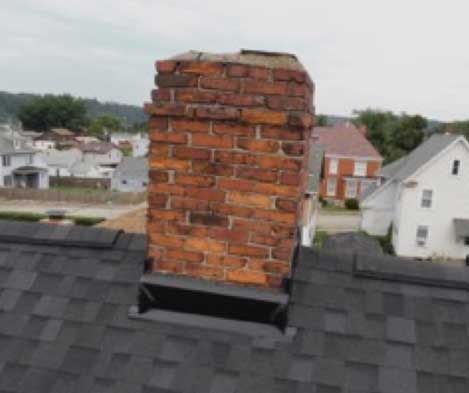 Brick chimney atop a dark-shingled roof with residential buildings in the background. The chimney bricks show signs of wear.