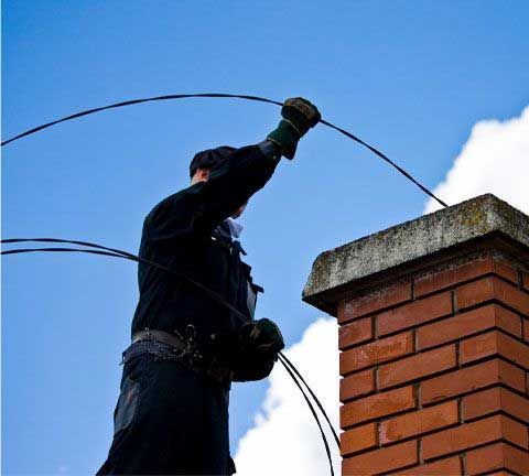 A chimney sweep, wearing black, cleans a brick chimney against a blue sky. They are reaching upward with a wire tool.