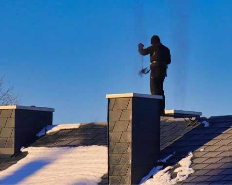 A chimney sweep on a rooftop in a clear blue sky. He is cleaning a chimney in winter with snow visible.