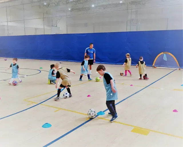 Children practicing soccer drills in a gym with cones and a coach watching