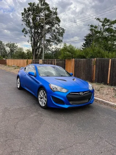 Blue sports car parked on a cracked asphalt road with a wooden fence and trees in the background.