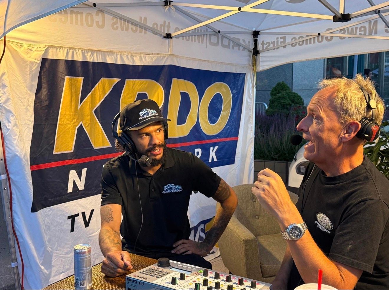 Two men broadcasting at an outdoor event under a branded tent, one speaking into a microphone.