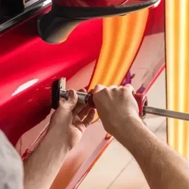 Hands using tools to remove a dent from a red car panel.