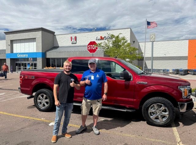 Two men by a red Ford F-150 truck in a store parking lot, one giving a thumbs up.