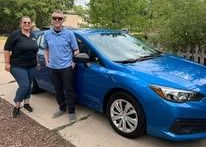Two people stand by a blue car; a man and woman smile. They are on a driveway in front of a house.