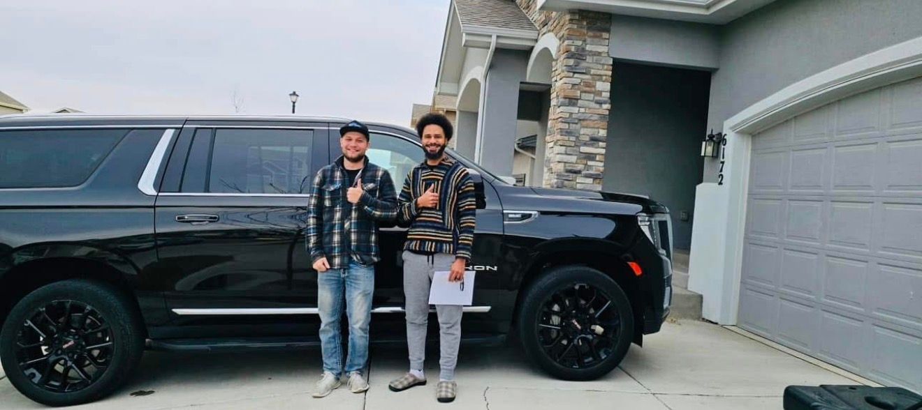 Two men give thumbs up next to a black SUV parked in front of a house with a gray exterior.
