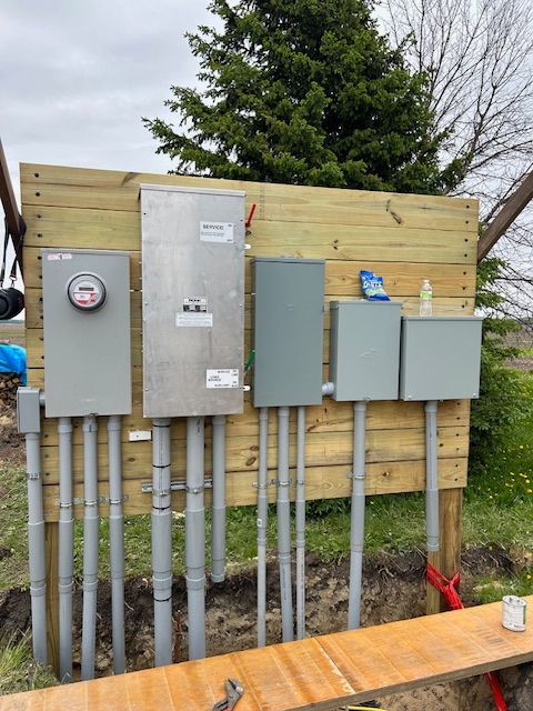 Electrical panel setup on a wooden board: meter, main panel, and subpanels, with gray conduits in a dirt trench.