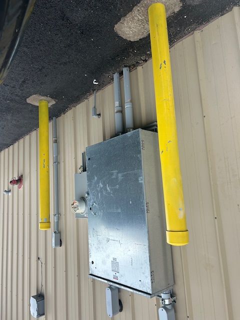 Yellow bollards protect an electrical box on a building's exterior. Gray conduits and beige siding are also visible.