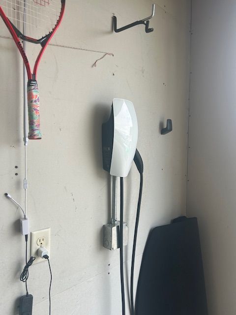 Wall-mounted EV charger next to a tennis racket and electrical outlet in a garage.