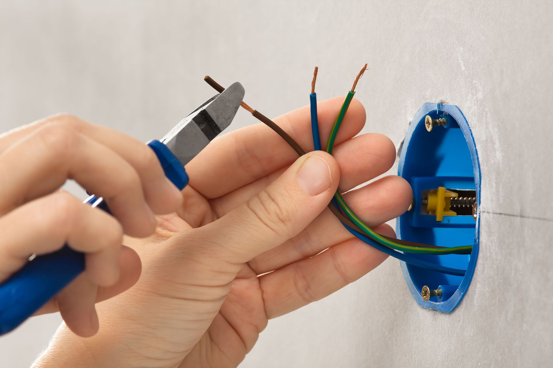 Person using blue handled pliers to strip a brown wire, next to a blue electrical box with exposed green and blue wires.