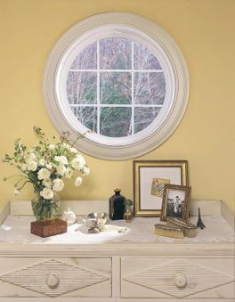 A round window is above a dresser with a vase of flowers on it.