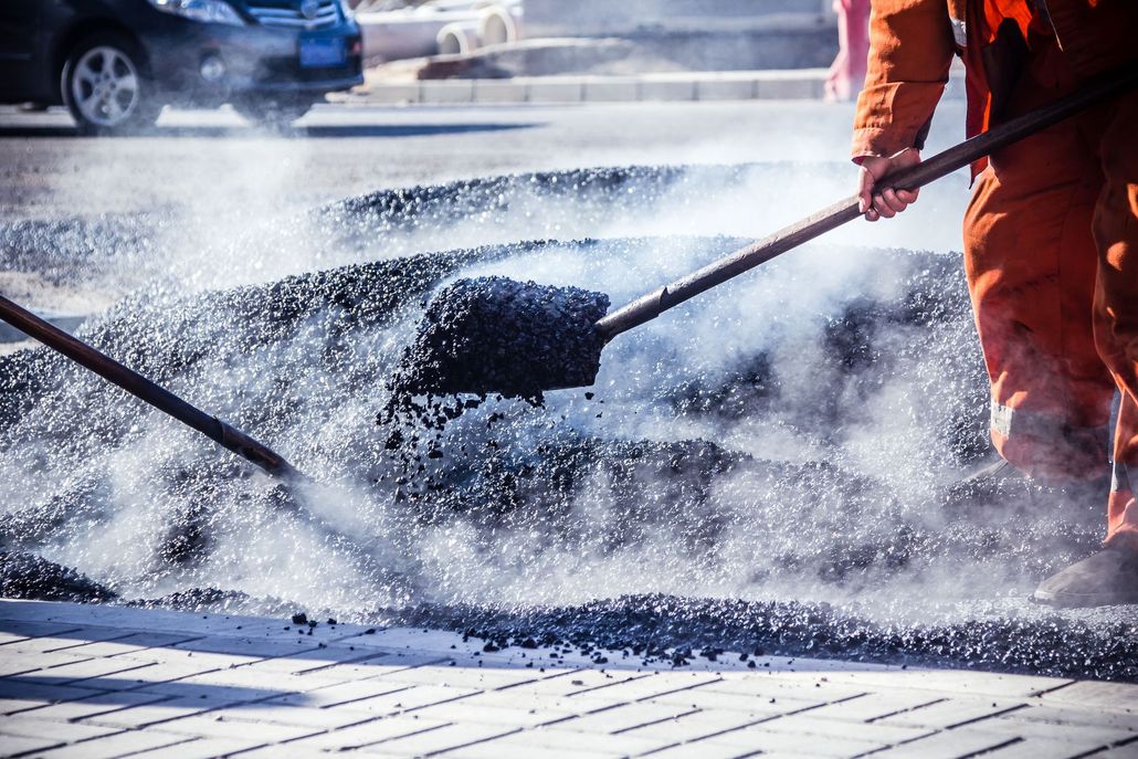 Worker spreading hot asphalt on a road with a shovel, with steam rising nearby