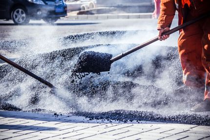 Worker spreading hot asphalt on a road with a shovel, with steam rising nearby