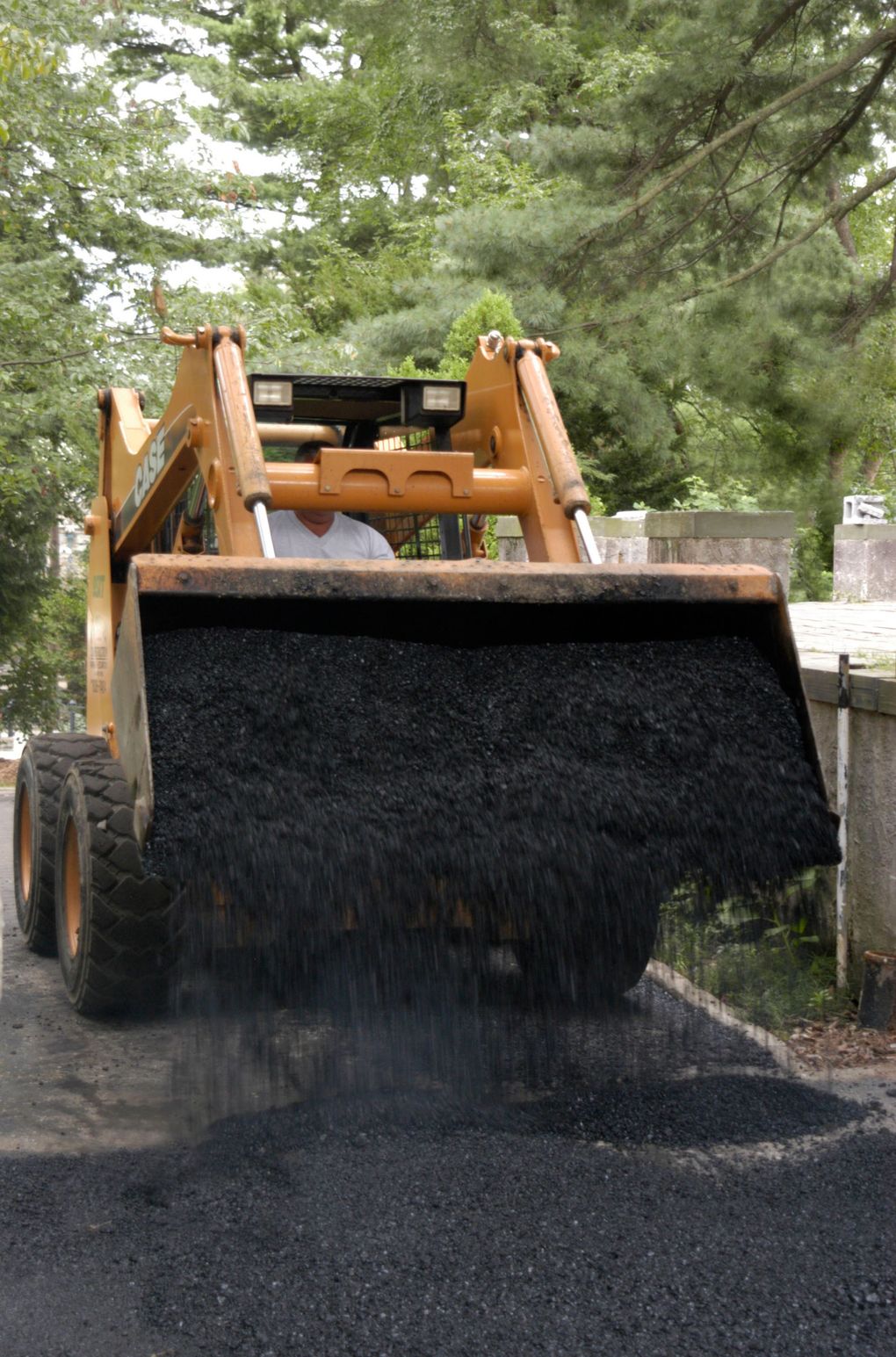 Front loader dumping black gravel onto a driveway beside a stone wall under trees