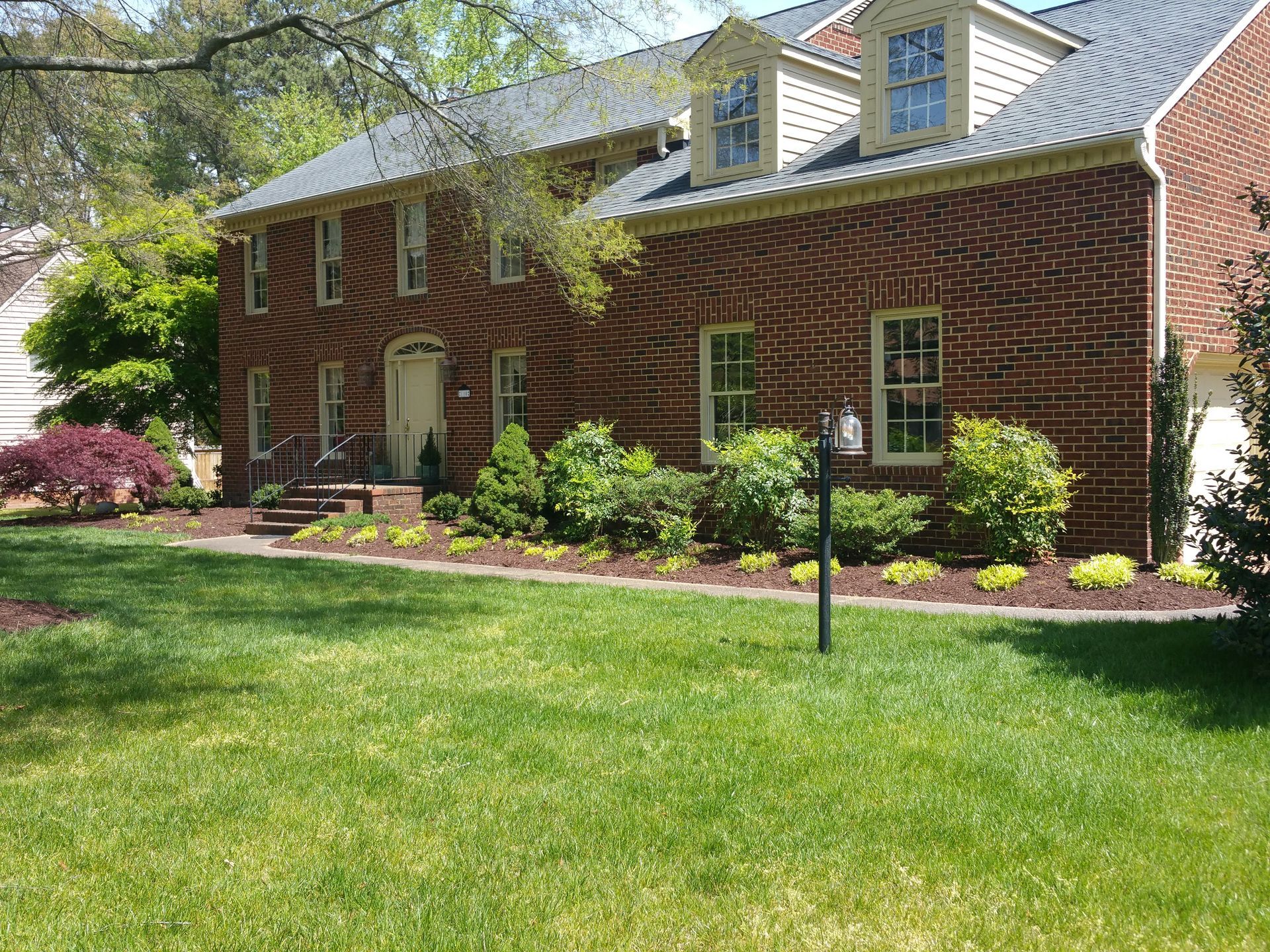 Brick house with white trim and dormer windows, front lawn, and landscaped shrubs in bright daylight