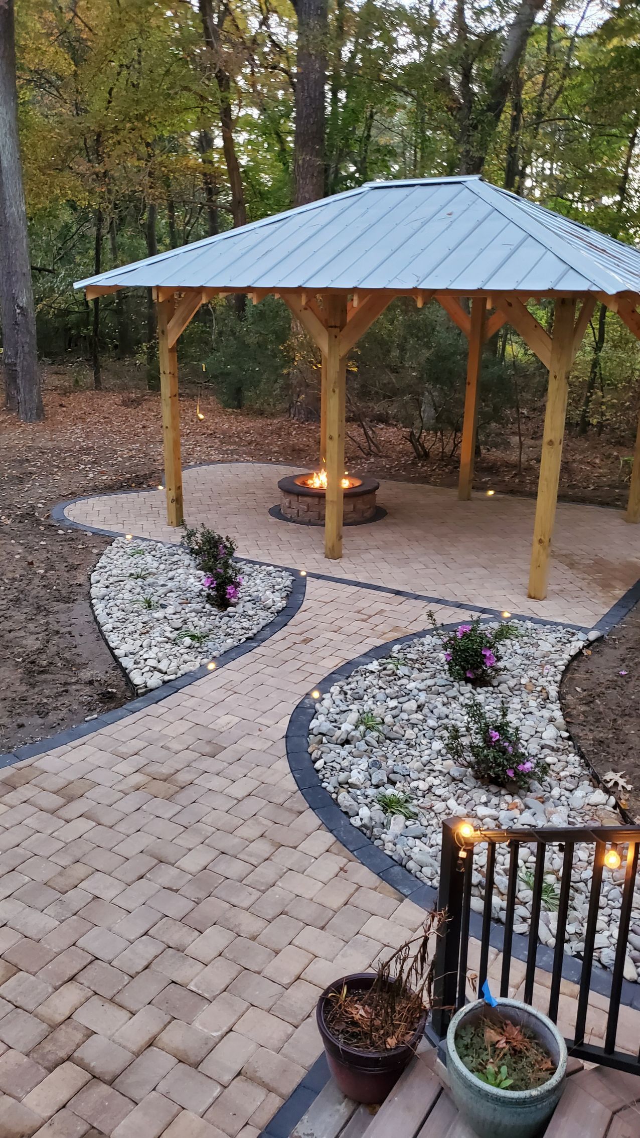 Covered patio with wooden gazebo, stone path, and landscaped garden beds in a wooded backyard.