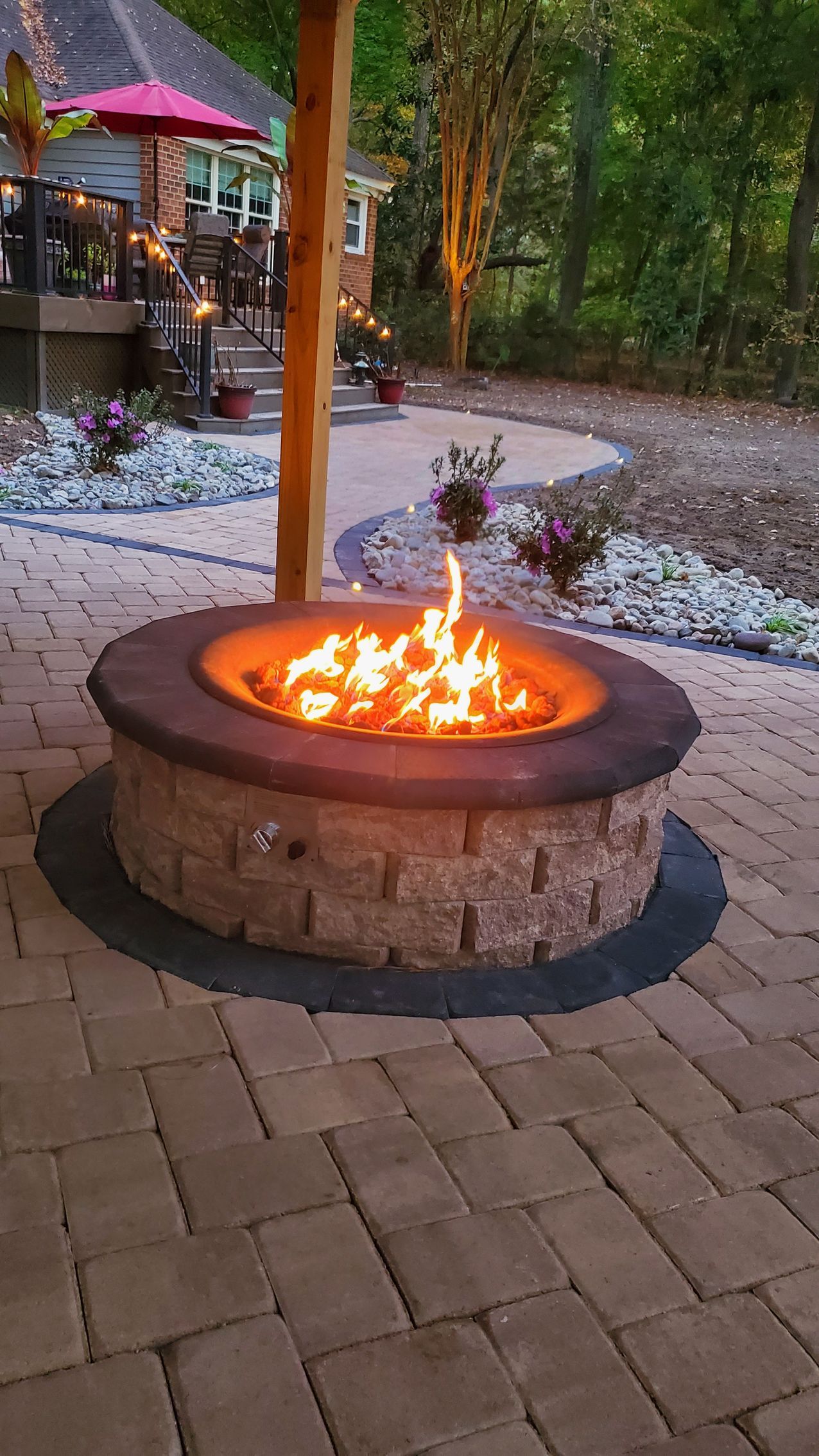 Stone fire pit with bright flames on a brick patio in a wooded outdoor setting at dusk