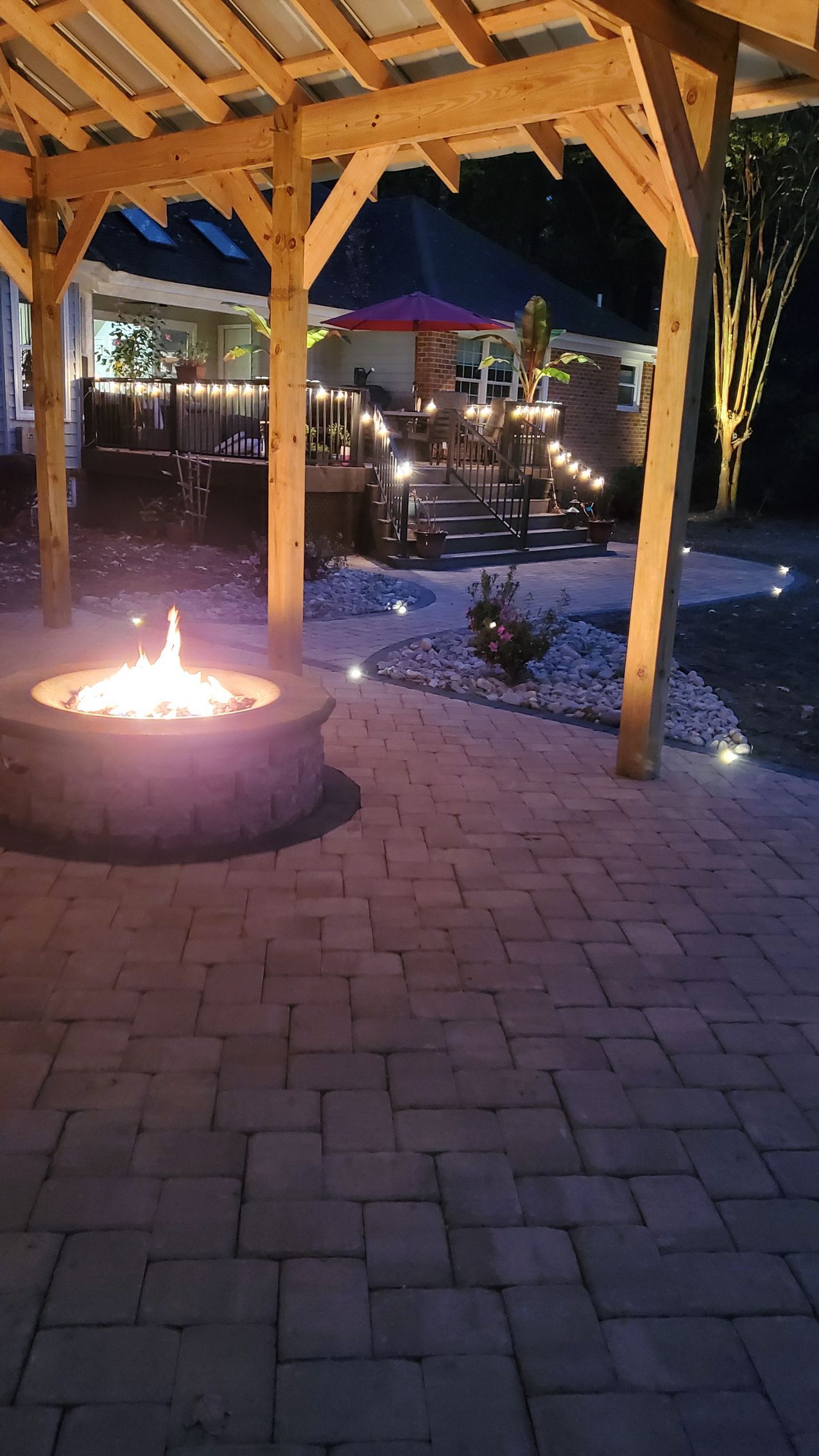 Fire pit under a wooden pavilion at night, with brick pavers and soft garden lights nearby.