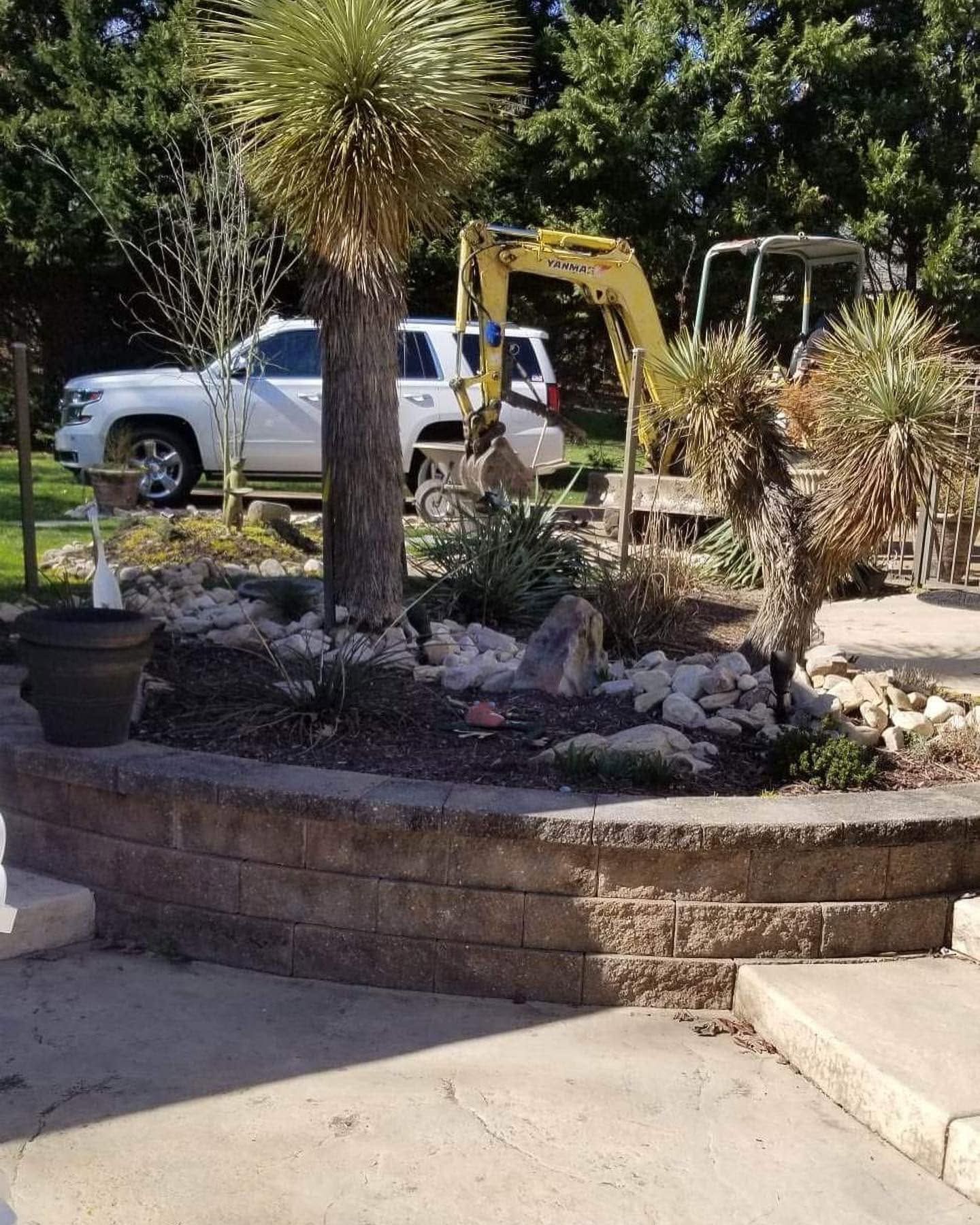 Yellow excavator beside a white pickup truck in a landscaped yard with yucca trees and stone planter walls