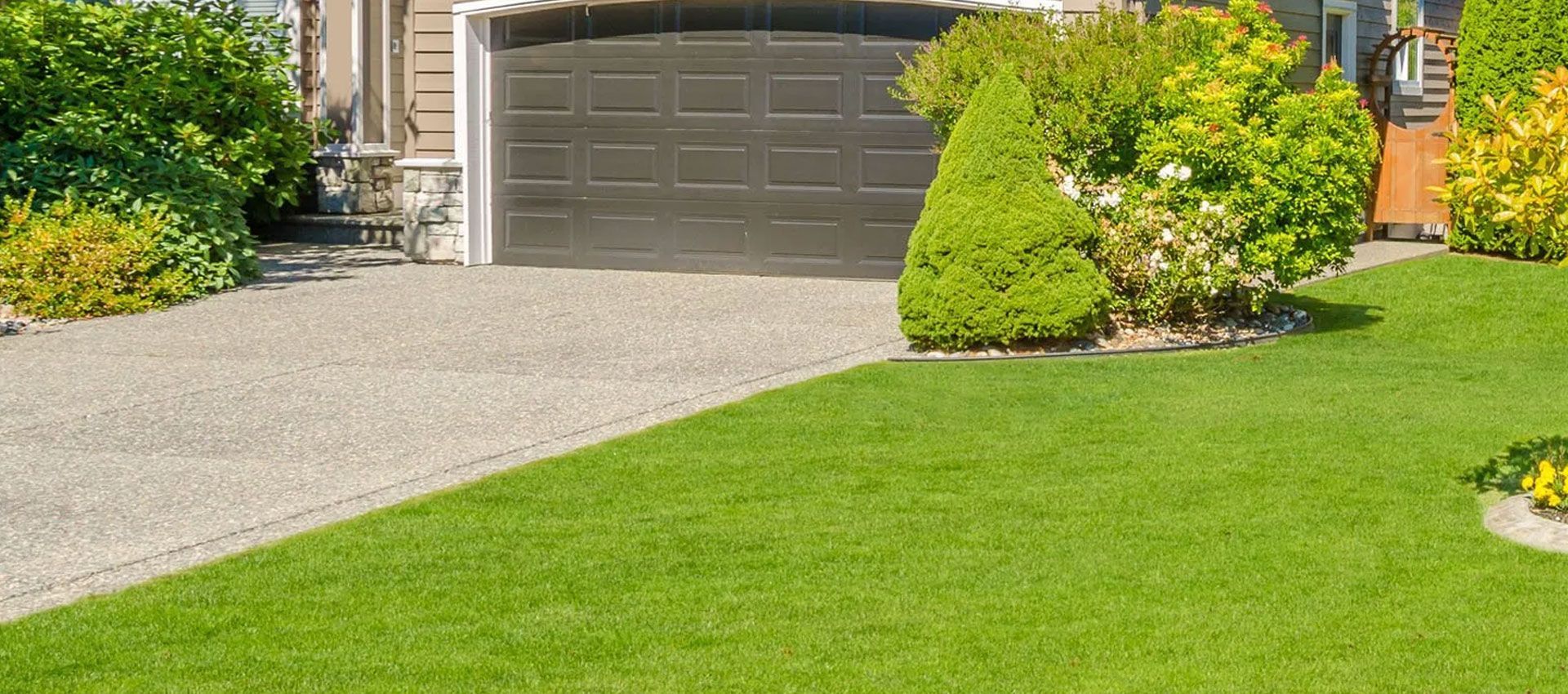 A brown garage door sits at the end of a gravel driveway, bordered by a green lawn, shrubs, and a small tree.