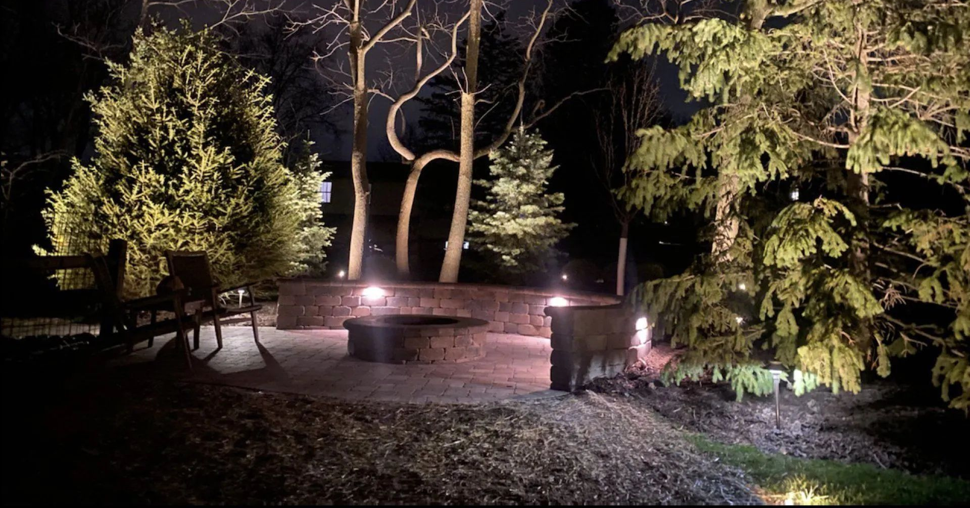 A stone fire pit and curved seating wall illuminated by outdoor lighting in a backyard at night, surrounded by trees.