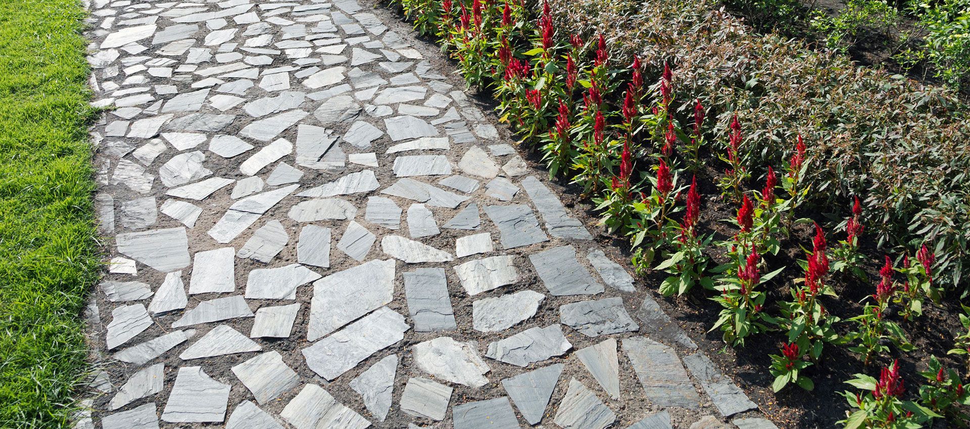 A stone path made of irregular gray pieces, bordered by a patch of green grass on one side and red flowers on the other.