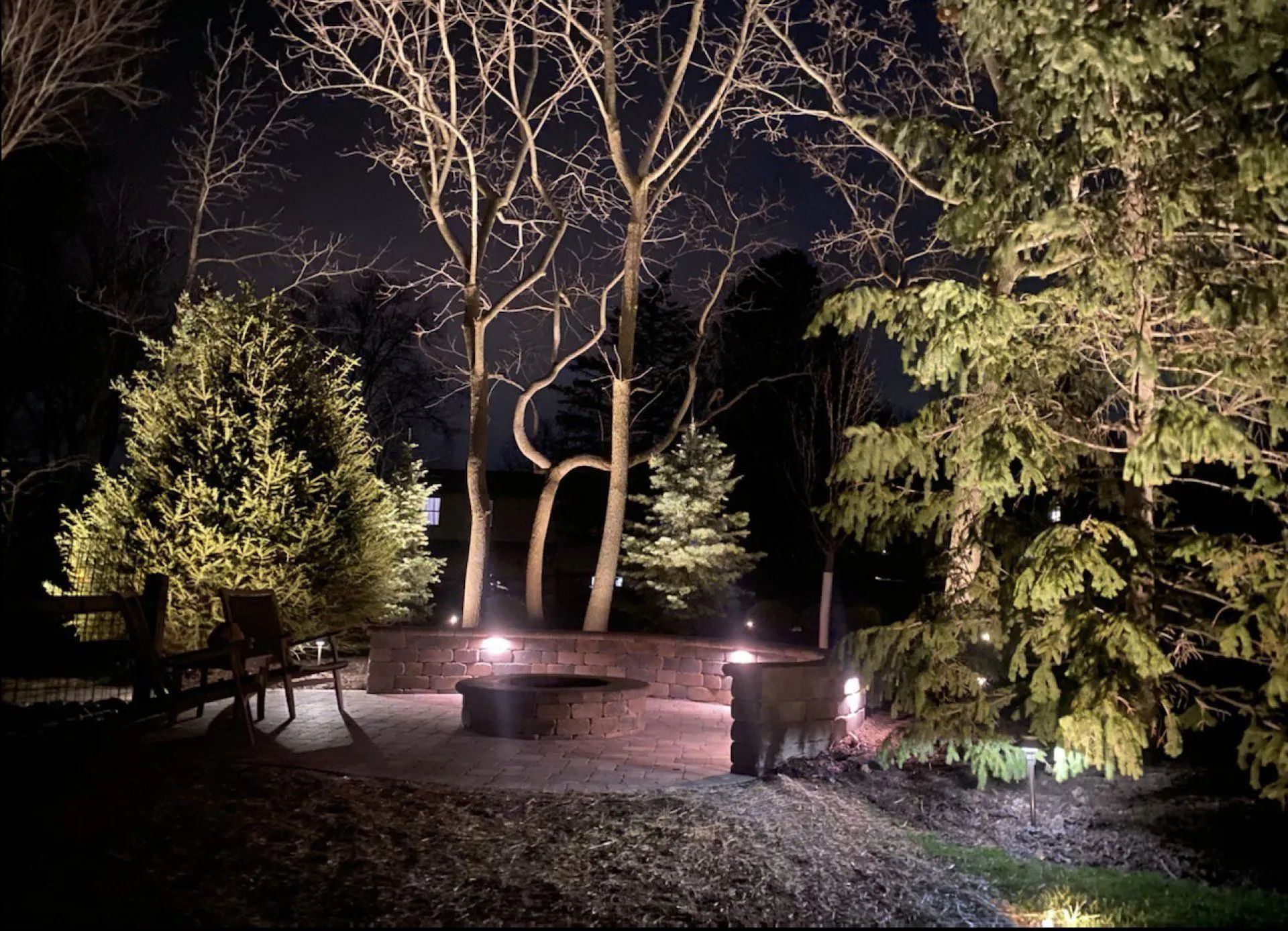 A backyard fire pit area at night, illuminated by landscape lighting, surrounded by trees and a stone wall.