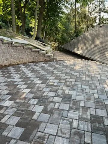 A patio with a multi-sized, gray-toned paver pattern next to a stone retaining wall and staircase, overlooking a roof.