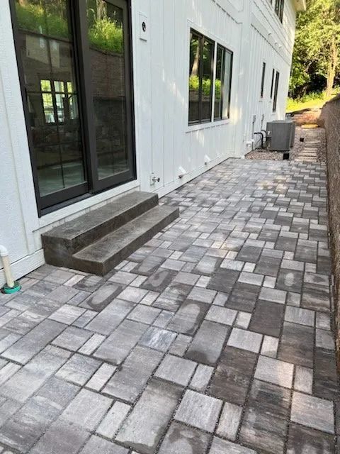 A light-colored house exterior with a gray stone paver patio and concrete steps leading to sliding glass doors.