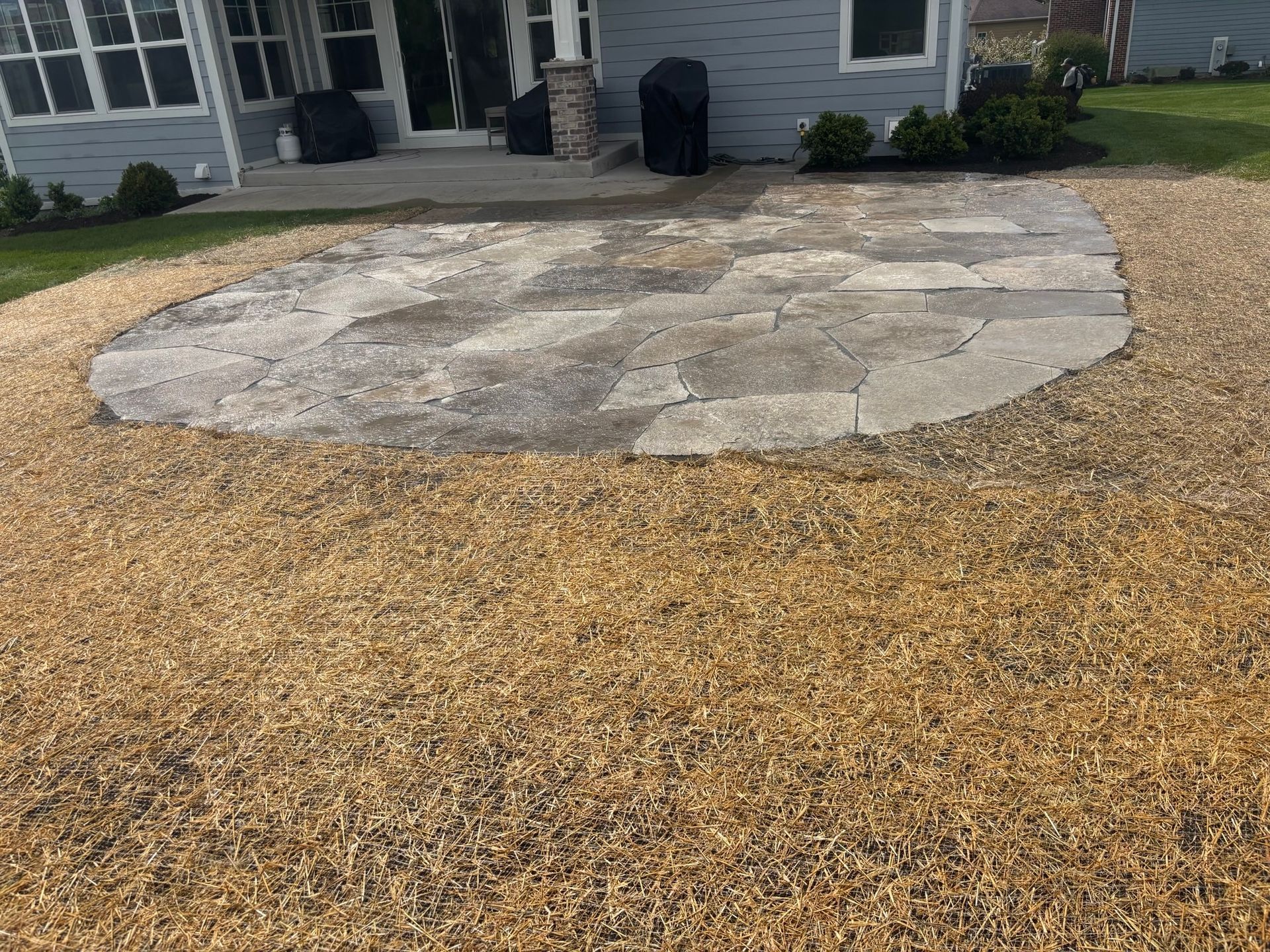 Small stone patio in a dry front yard beside a house with light gray siding.