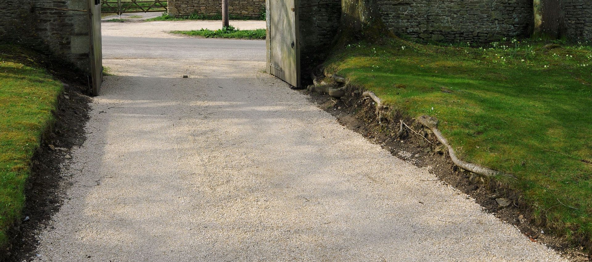 A gravel path leads through an open stone gate into a garden, flanked by green grass and dark soil borders.