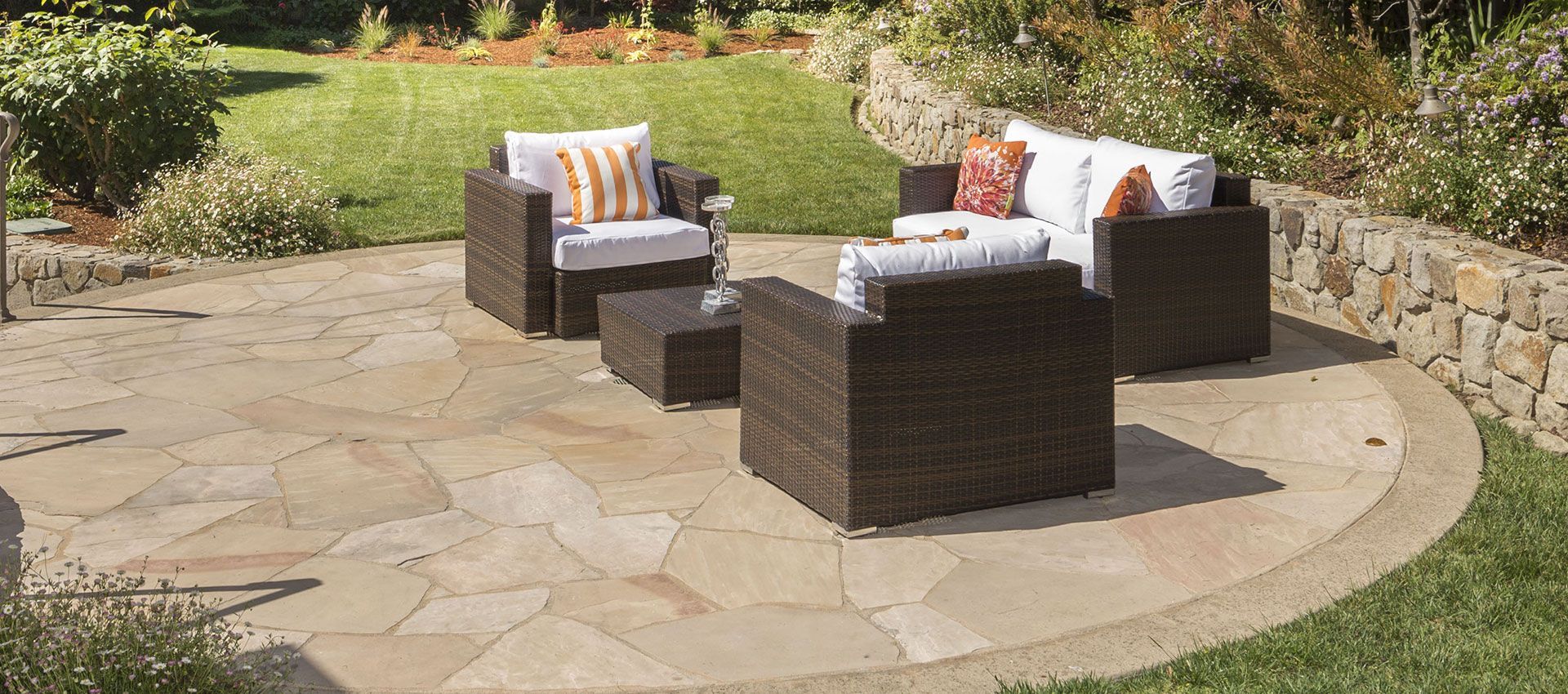Wicker outdoor furniture set with white cushions on a circular stone patio in a backyard.